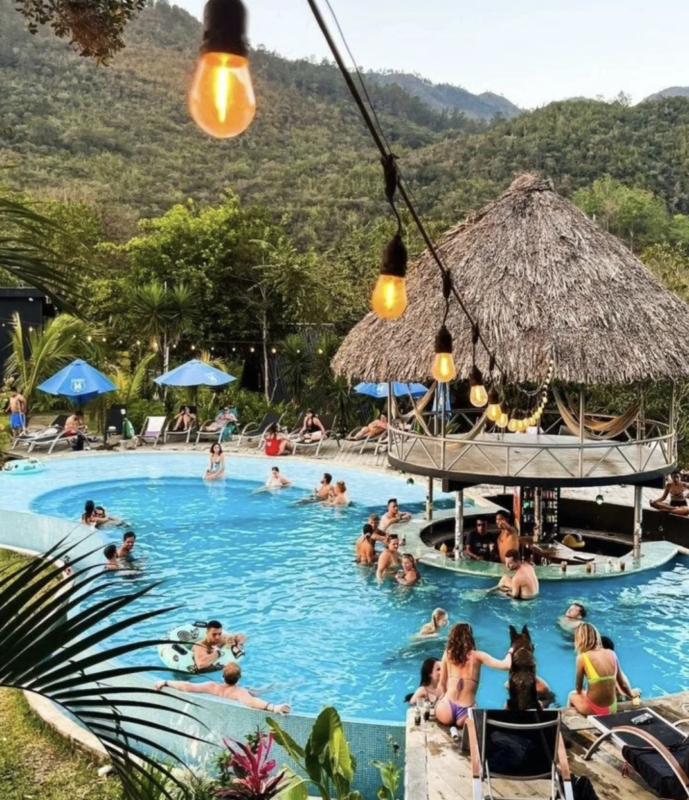 Sunny resort pool area with string lights, a thatched-roof bar, and people swimming and lounging.