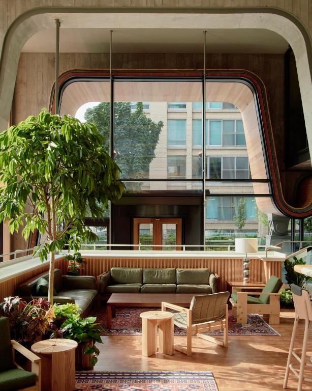 Indoor lounge with green leather seating, wooden chairs and tables, plus a large potted tree; curved architectural window behind.