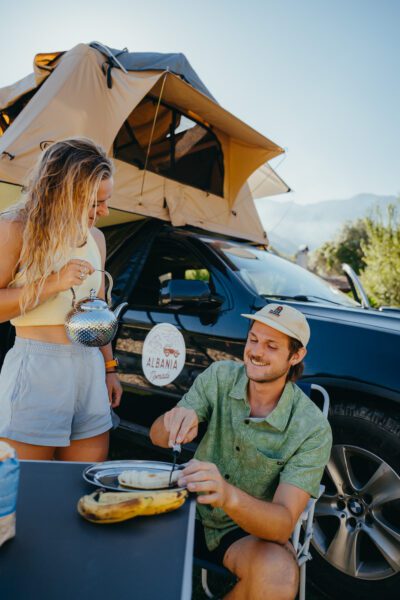 A woman in a yellow top pours tea from a shiny kettle while a man in a green shirt sits at a table outside a car with a rooftop tent behind them.