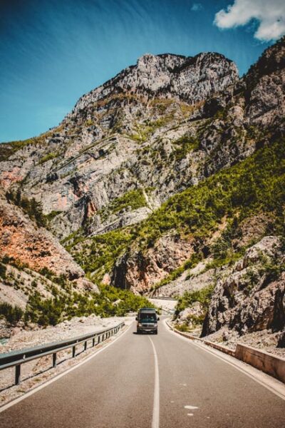 Black SUV driving away on a winding two-lane mountain road, flanked by rugged rocky cliffs and green vegetation beneath a clear blue sky.