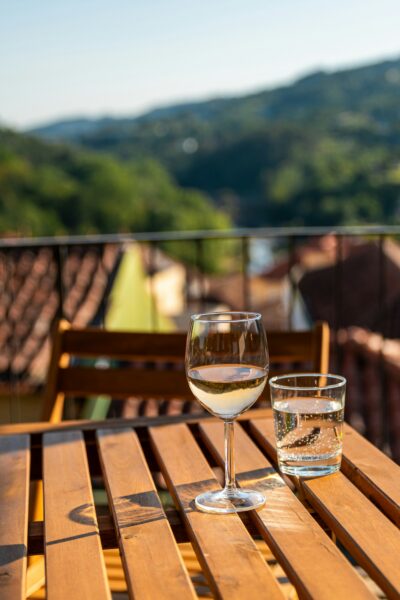 Wine glass and a glass of water on a sunlit wooden patio table with blurred hills in the background.