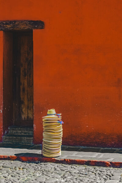 Tall stack of straw hats with a hat on top, leaning against an orange wall beside a doorway on a cobblestone sidewalk.