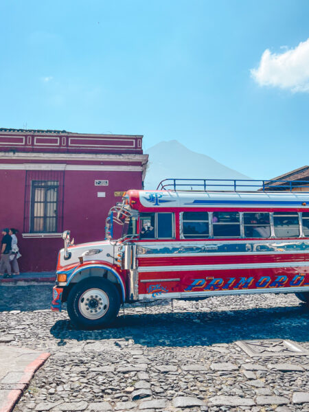 Colorful bus parked on a cobblestone street with a red building behind and a mountain in the distance under a blue sky.