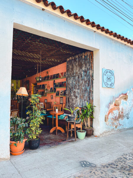 Open-air cafe with wooden furniture and potted plants along a blue storefront doorway.