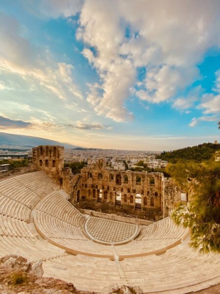 Ancient stone amphitheater with curved rows of seating and ruined arches, overlooking a city under a blue sky with clouds.