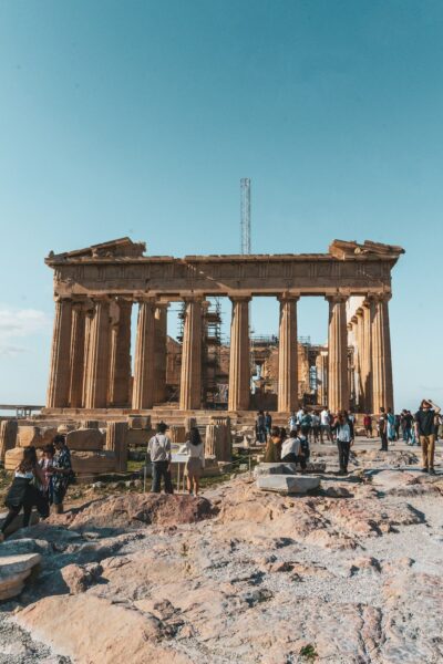 Ancient Parthenon ruin with visitors exploring the rocky foreground under a clear blue sky, Athens, Greece.