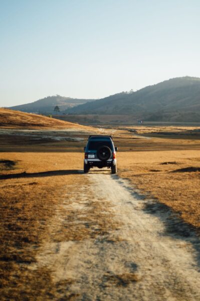 Blue SUV driving away on a dusty dirt road through a dry, golden field with hills in the distance.