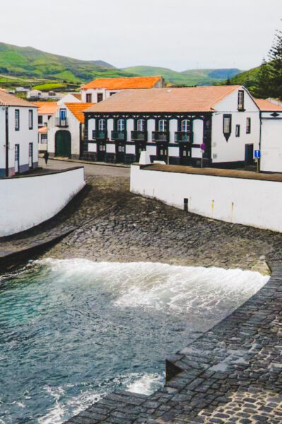 Quaint harbor town with white buildings, red-tiled roofs, and a curved stone promenade along the sea; green hills in the distance.