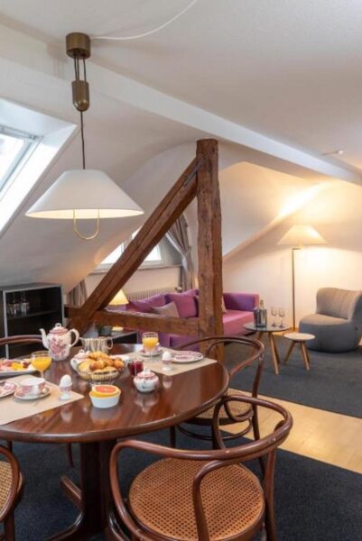 Cozy attic dining area with a round wooden table set for breakfast, featuring a teapot, pastries and juice near a rustic wooden beam and purple seating in the background toward a lounge area.] } } Wait that's bad. I must provide proper JSON. Let's craft proper array strings. Remove stray brackets. Let's deliver: