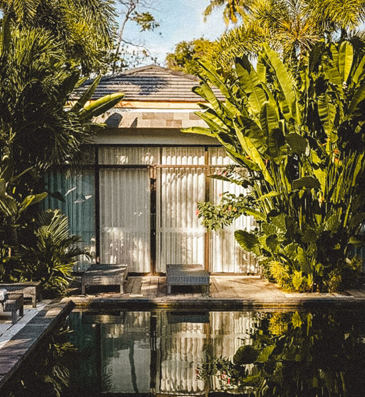 Small bungalow surrounded by lush tropical plants with a rectangular pool in the foreground.