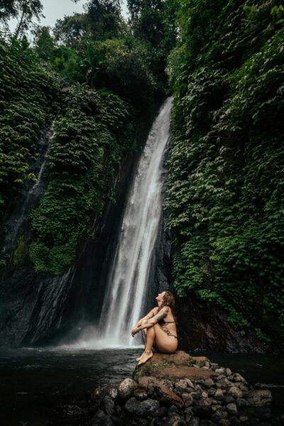 Person in a bikini sits on rocks at the base of a tall jungle waterfall, surrounded by lush green foliage and misty pool water