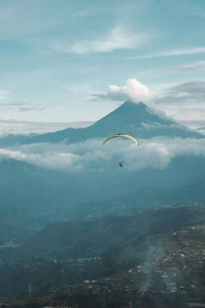 Paraglider soaring above misty mountains with a tall volcano in the background.