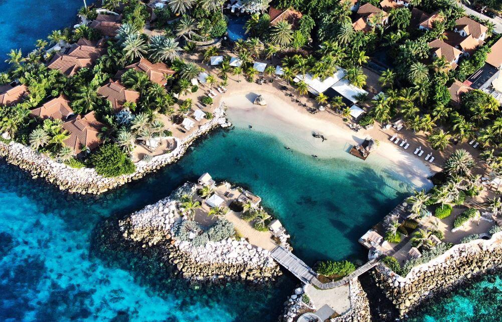 Aerial view of a tropical resort with turquoise water, white sand, palm trees, and villas along the shore.