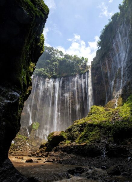 Waterfall in a deep, mossy canyon with cliffs on both sides and a rocky pool at the base under a blue sky.