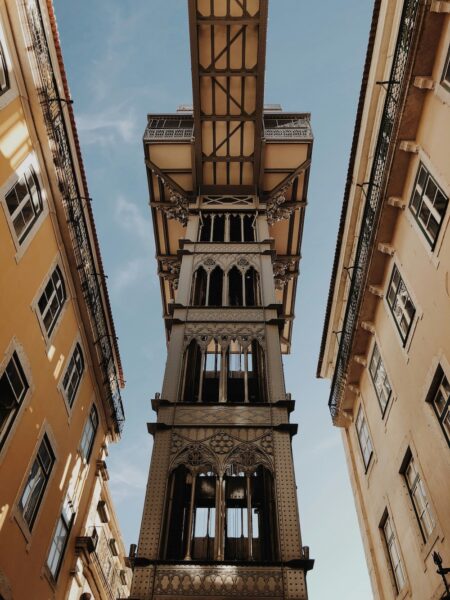 Santa Justa Lift, a tall ornate iron tower rising between surrounding orange/cream buildings against a blue sky.