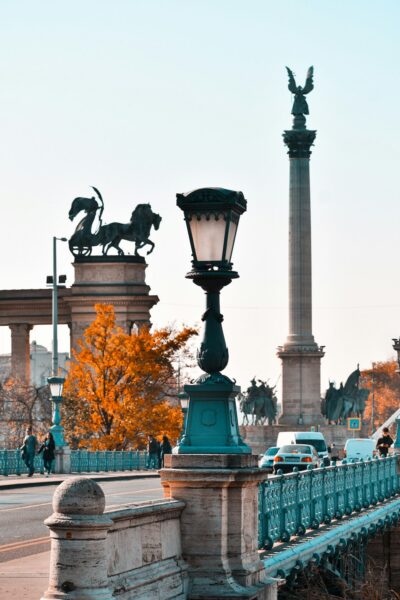 Ornate teal street lamp on a stone bridge with orange autumn trees and a tall column statue in the background