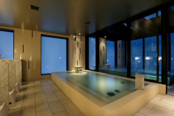 Indoor spa with a rectangular beige-tiled hot tub, dim lighting, and floor-to-ceiling windows along two walls at night.