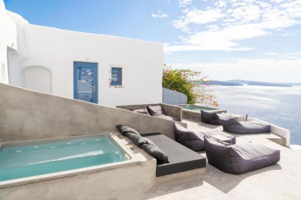 Sunny white terrace with a blue door and panoramic sea view; cozy gray lounge seating and a rectangular hot tub.