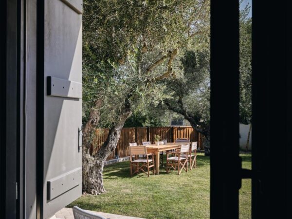 Open doorway looks out onto a sunlit backyard dining area with a wooden table and chairs under olive trees.
