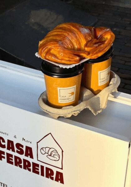 Two takeaway coffee cups with black lids and orange sleeves sit in a cardboard carrier, a glossy circular pastry resting on a napkin atop them, labeled Casa Ferreira.