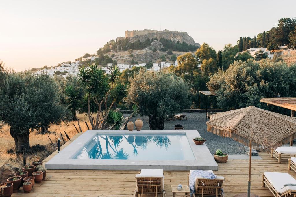 Outdoor villa with a rectangular pool on a wooden deck, olive trees, and a hillside fortress in the distance at sunset.