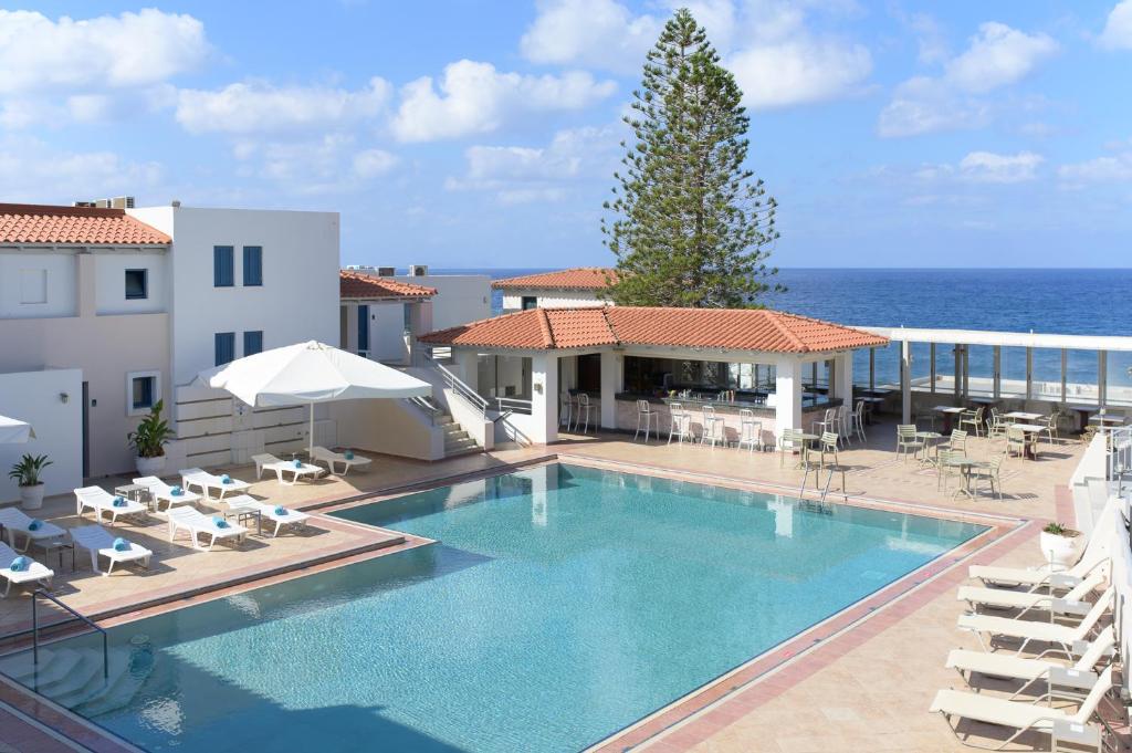 Resort pool area overlooking the ocean with white buildings, sun loungers, umbrellas, and a shaded bar area.
