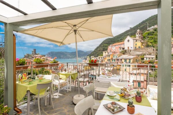 Terrace cafe with a large umbrella, set tables, and a coastal hillside town in the background.