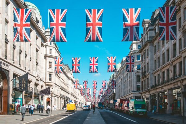 City street lined with Union Jack flags overhead, upscale storefronts, pedestrians, and vehicles present.