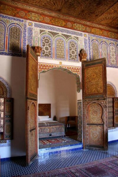 Ornate Moroccan-style bedroom alcove with carved wooden doors opened, revealing a bed and small table.