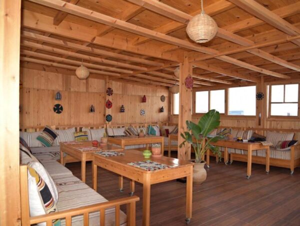 Rustic wooden lounge with beige sectional couches, colorful cushions, and square coffee tables under exposed beams.