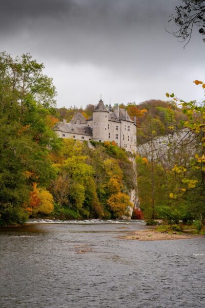 Stone castle perched on a cliff above a winding river, surrounded by colorful autumn trees under a cloudy sky.