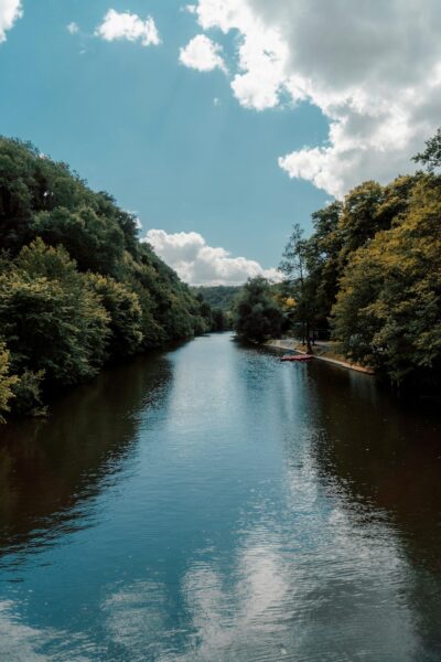 River flowing between tree-lined banks under a blue sky with fluffy clouds; a small dock with kayaks on the right shore.