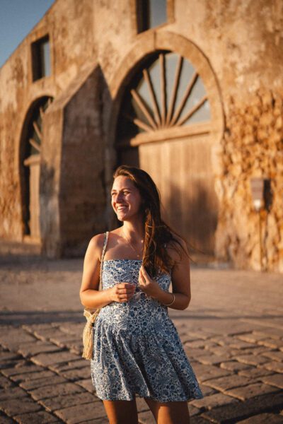 A smiling woman in a blue floral sundress stands on a cobblestone street near an old stone archway at sunset.