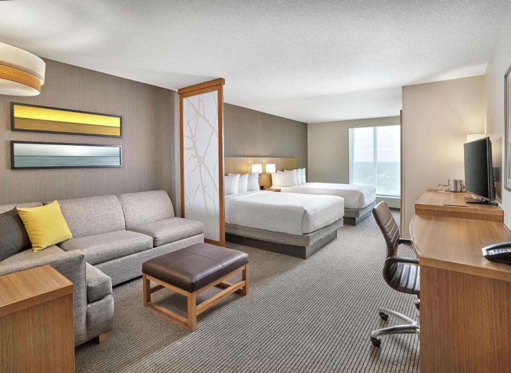 Hotel suite: living area with gray sofa, yellow pillow, ottoman, and two beds in the background.