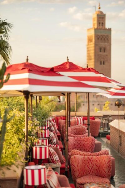 Rooftop lounge with red-and-white striped umbrellas and pink patterned chairs along a water feature, with a tall stone tower in the distance.