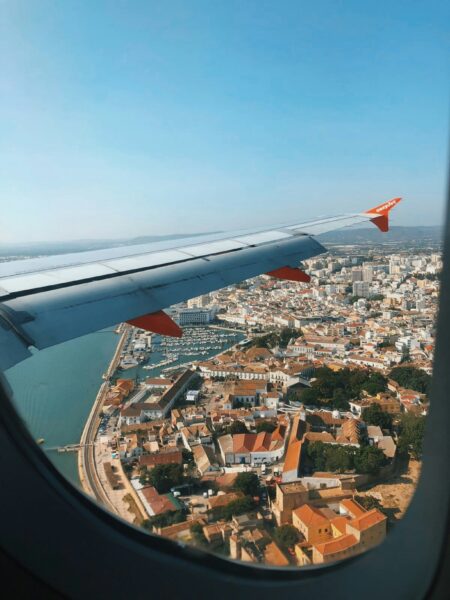 Airplane wing with easyJet logo viewed from window over a coastal town with a marina and red-tiled roofs beneath.