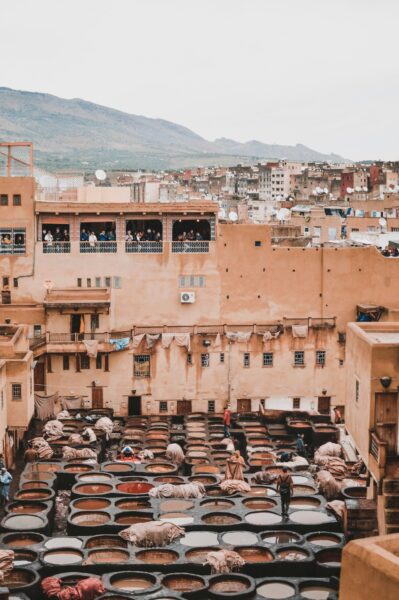 Moroccan open-air tannery with rows of circular dye vats and workers among beige buildings. Nearby spectators overlook from a rooftop.