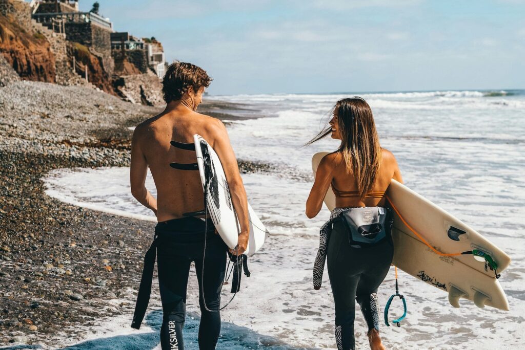 Couple walking along a pebbly beach with surfboards toward the ocean, rocky cliffs in the background.