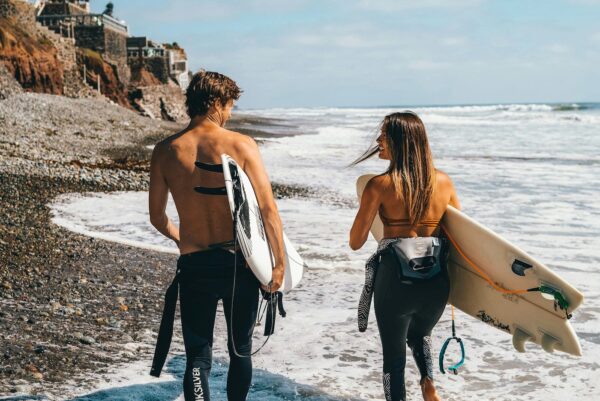 Couple walking along a pebbly beach with surfboards toward the ocean, rocky cliffs in the background.