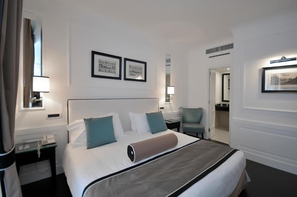 Modern hotel room with a king bed, white linens, teal pillows, and a cylindrical bolster at the foot. Two nightstands and wall lamps flank the bed.