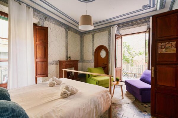 A cozy hotel bedroom with a white bed and rolled towels, wooden headboard, a green chair and a purple sofa near a balcony door.