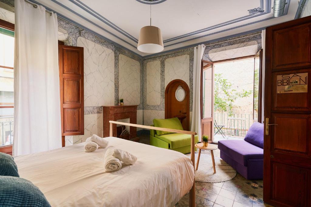 A cozy hotel bedroom with a white bed and rolled towels, wooden headboard, a green chair and a purple sofa near a balcony door.