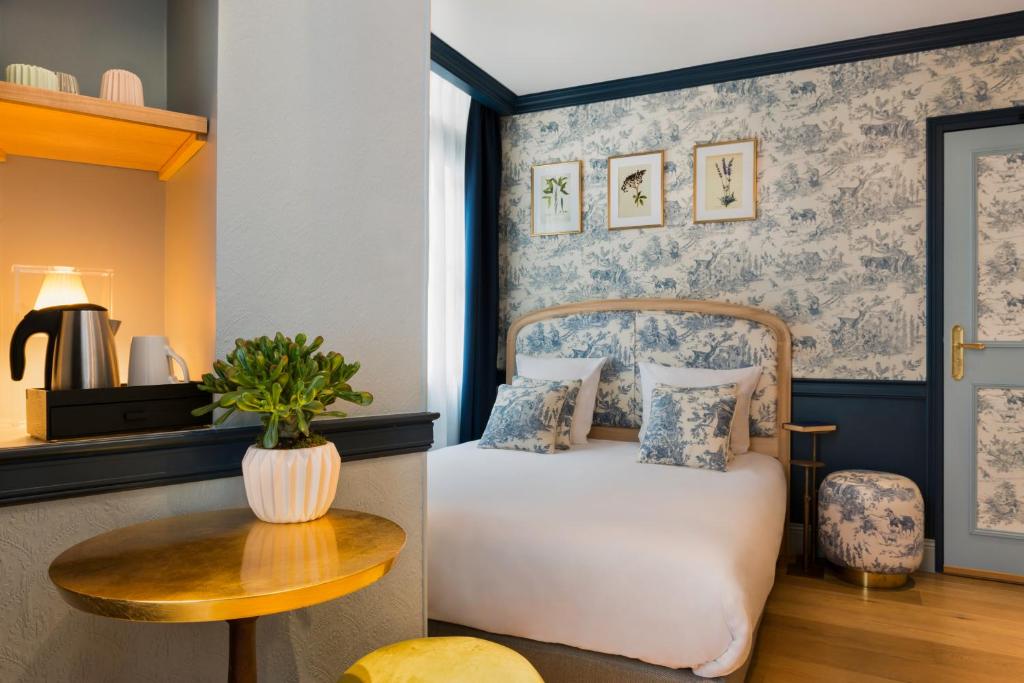 Hotel room with a blue toile wallpaper, bed with white linens and matching pillows, and a small round wooden table with a plant. A kitchenette area with kettle and cups is visible on the left.