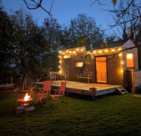 Cozy wooden cabin at dusk with warm string lights along the deck, outdoor chairs, and a glowing fire pit on the lawn nearby