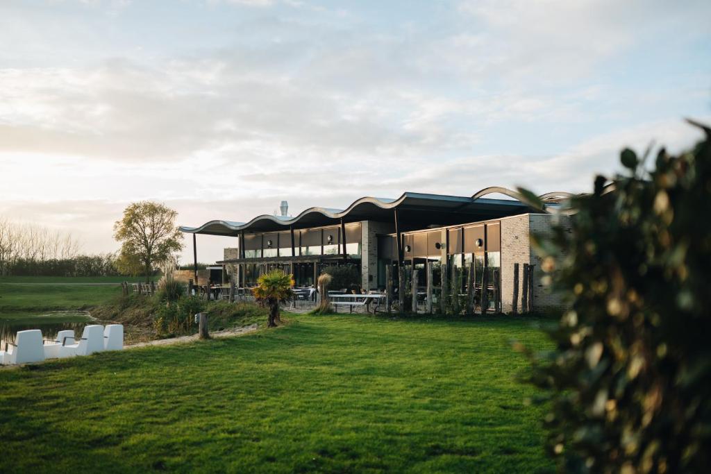 Modern café with a wavy roof, glass walls, and outdoor seating beside a green lawn and small water feature at sunset.