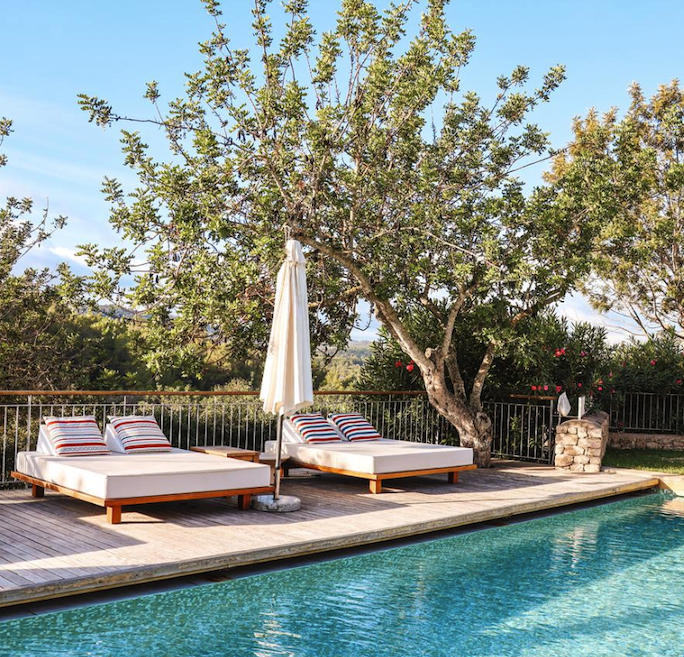 Two white cushioned lounge beds with striped pillows on a wooden pool deck, a closed umbrella between them beside a blue pool under a tree.