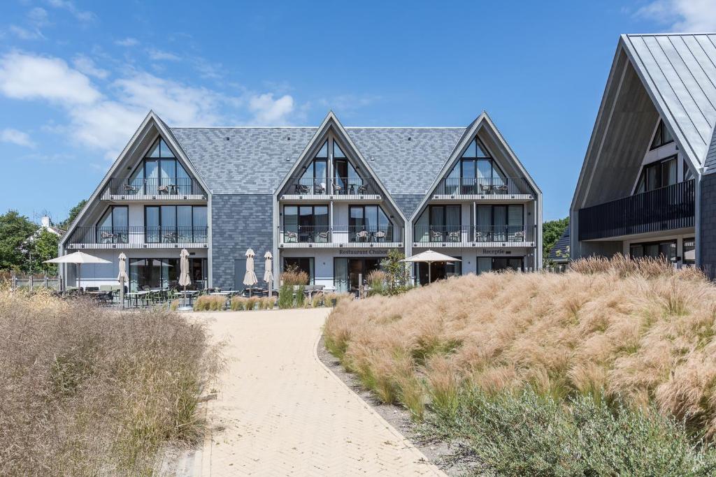 Row of modern A-frame hotel buildings with balconies, facing a sandy path and tall beach grasses.