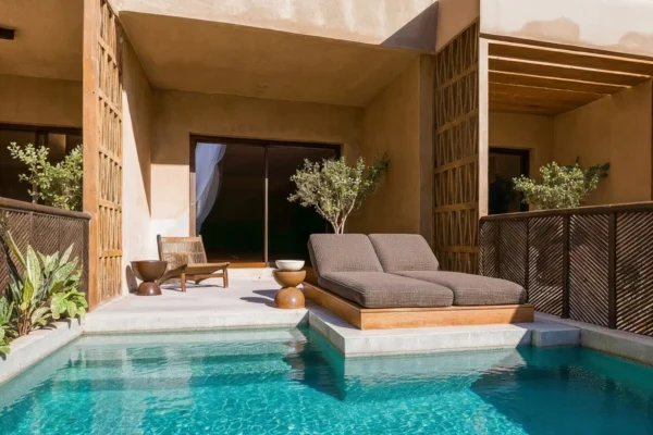 Outdoor poolside patio with two brown cushioned lounge chairs, a small wooden table, and potted trees beside a beige stucco building.