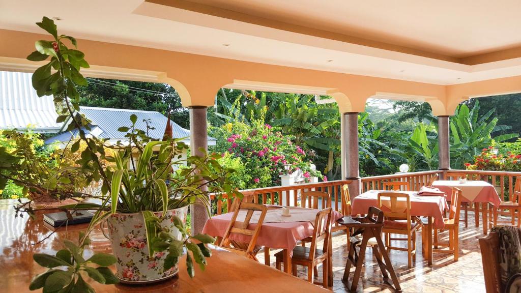 Open-air veranda dining area with pink tablecloths, wooden tables and chairs, and a large potted plant in the foreground; lush tropical garden in the background.