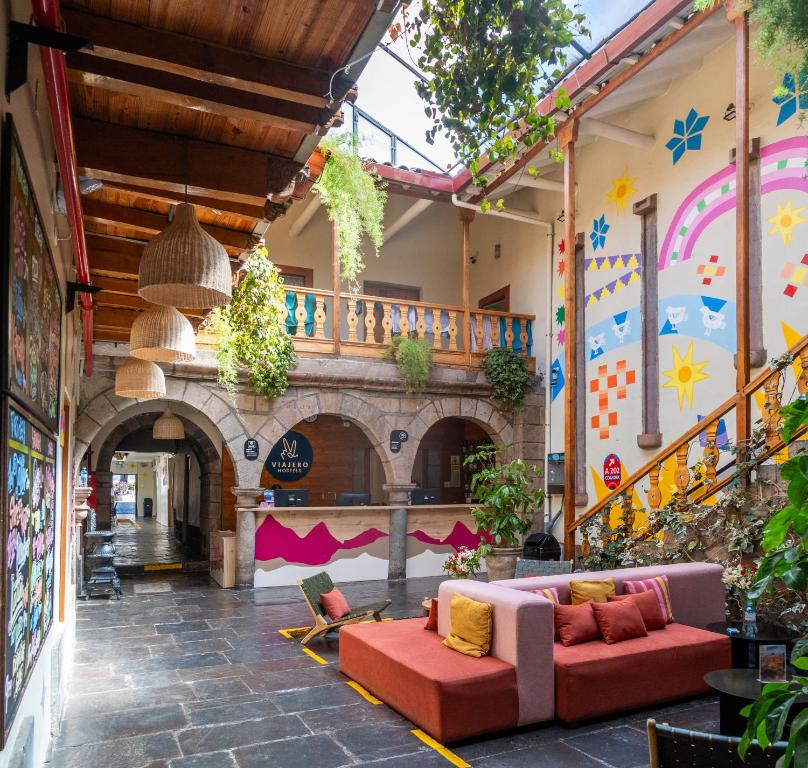 Colorful hotel courtyard with stone arches, wooden balcony, hanging plants, and a bright mural; plush orange seating in the center.